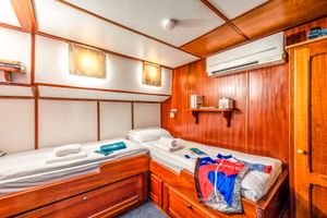 Interieur van MS Caprice hut met twee bedden, houten lambrisering, airconditioning, wandlampen en laden. Handdoeken en kleding op bedden.