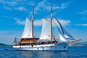 MS Macek, een wit zeilschip met twee masten en witte zeilen, varend op kalm blauw water onder een heldere hemel met verspreide wolken.