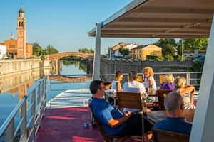 Passagiers ontspannen op het zonnedek van MS Ave Maria in Portegrandi, met kanaal, brug en kerktoren op de achtergrond.