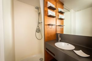 Compacte badkamer op MS Ave Maria met witte douche, ovale wastafel in donker blad, houten wandpanelen, planken en spiegel.