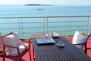 Zonnedek met houten tafel, stoelen met witte kussens en rode vloer met uitzicht op de Venetiaanse lagune met verre eilanden.