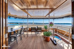 Overdekt buitendek van MY Olmissum met houten plafond, eettafel, stoelen en panoramisch uitzicht op het meer door glazen panelen.