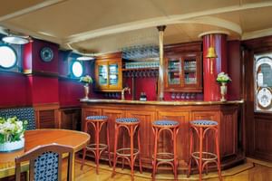 Houten bar met vier rode barkrukken op schip Mare Frisium. Rode wanden, patrijspoorten, hangende glazen en bloemen creëren gezellige sfeer.