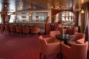 Elegante bar lounge op MS Switzerland met koraalkleurige fauteuils, ronde tafels, rood tapijt en houten plafond met decoratieve verlichting.