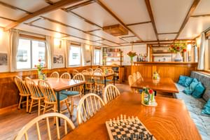 Lichte salon aan boord van MS Flora met houten plafondbalken, eettafels met Windsor-stoelen, schaakspel, ingebouwde bar en blauwe kussens.