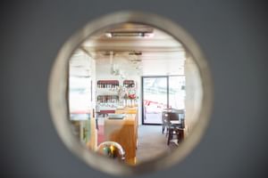 Zicht door ronde patrijspoort op helder interieur van MS Ave Maria met houten balie, planken, eethoek en glazen toegangsdeur.