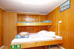 Houten betimmerde hut op Kazimir schip met stapelbed en eenpersoonsbed, wit beddengoed, handdoeken en ingelijste kustfoto.