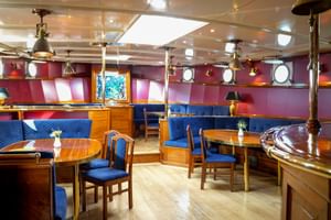 Interieur van Mare Frisium scheepssalon met houten tafels, blauw gestoffeerde zitplaatsen, koperen pilaren, hanglampen en ronde patrijspoorten op rode wanden.
