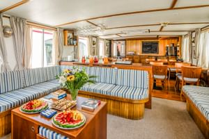 Interieur van MS Caprice salon met blauw-wit gestreepte gestoffeerde zitplaatsen, houten meubels, fruitschalen op salontafel en kombuis op de achtergrond.