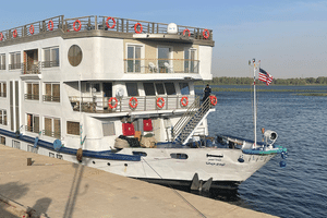 Wit Nebu riviercruiseschip met blauwe rand aangemeerd aan de Nijl. Het schip heeft meerdere dekken met balkons en een Amerikaanse vlag achteraan.
