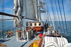 Dek van zeilschip Mare Frisium met witte zeilen, tuigage, houten banken en stuurhut. Blauwe lucht en zee op de achtergrond.