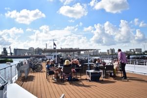 Passagiers ontspannen op het houten zonnedek van DE Willemstad met tafels, stoelen en stadsgezicht over het water onder blauwe lucht.