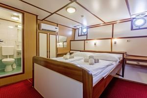 MS Amore benedendek hut met tweepersoonsbed, wit beddengoed, houten lambrisering, rood tapijt en badkamer met douche zichtbaar door open deur.