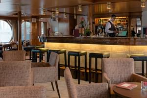 Moderne loungebar aan boord van MS Veloria met beige gestoffeerde stoelen, barkrukken, verlichte bar en grote ronde ramen met uitzicht op het water.