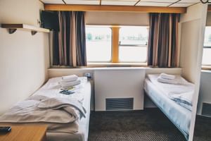 Interieur van een scheepshut met twee eenpersoonsbedden, wit beddengoed, handdoeken, gestreepte gordijnen en patrijspoorten met waterzicht in Willemstad.