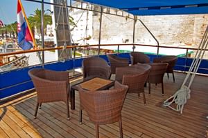 Houten dekterrasse van MS Macek met bruine rieten stoelen en tafels onder wit zonnescherm. Kroatische vlag zichtbaar, historische stenen muren op achtergrond.