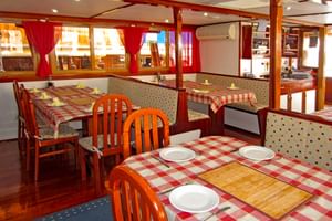 Interieur van MS Macek restaurant met houten tafels bedekt met rood geruite tafelkleden, oranje houten stoelen en rode gordijnen.