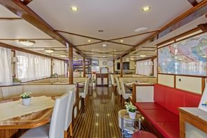 Interieur van MS Amore scheepssalon met witte eettafels, rode en beige zitplaatsen, houten plafondbalken en zeekaarten aan de wanden.