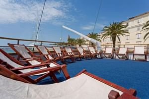 Zonnedek van cruiseschip MS AMORE met rijen houten ligstoelen met witte kussens op blauw dek, mediterrane gebouwen zichtbaar op achtergrond.