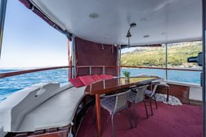 Overdekt buitendek van MS Mirabela met witte zitplaatsen, houten tafel, rieten stoelen en rood tapijt. Kroatische kust zichtbaar door ramen.