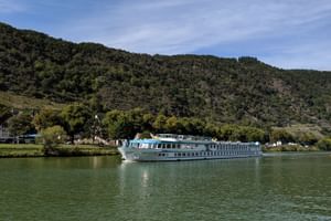 MS Switserland riviercruiseschip met witte romp en blauw bovendek varend op kalm groen water met beboste heuvels en dorp op de achtergrond.