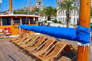 Zonnedek van MS Macek met houten ligstoelen op rijen onder blauw zonnescherm. Kroatische kuststad met palmbomen en historische gebouwen zichtbaar.