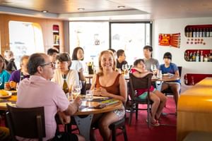 Interieur van MS Ave Maria salon restaurant met gasten aan tafels met wijnglazen. Grote ramen en decoratieve wanddisplays zichtbaar.