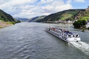 Riviercruiseschip MS Veloria varend op een brede rivier tussen groene heuvels met wijngaarden en kleine dorpen onder een gedeeltelijk bewolkte hemel.