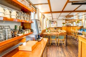 Interieur van MS Flora scheepssalon met houten balken, eettafels met stoelen, bar en planken met glaswerk en kopjes.