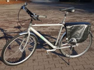 Witte Bird toerfiets met rechtop stuur, zwart zadel en grijze fietstassen op bagagedrager, geparkeerd op klinkers.