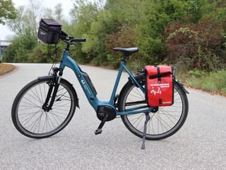 Blauwe e-bike met zwarte stuurtas en rode achtertassen geparkeerd op geasfalteerd pad omringd door groene bomen en struiken.