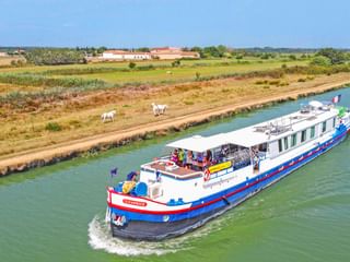 MS Caprice riviercruiseschip met blauwe en rode strepen varend op een kanaal. Landelijk landschap met velden en witte paarden op de oever.