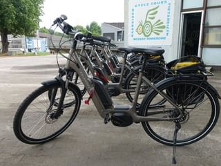 Rij grijze unisex e-bikes met bagagedragers geparkeerd bij een fietsverhuurstation. De fietsen hebben middenmotoren en standaards.
