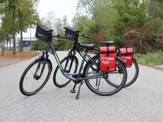 Twee zwarte toerfietsen met stuurtas en rode achtertassen geparkeerd op een verhard pad omringd door bomen en herfstbladeren.