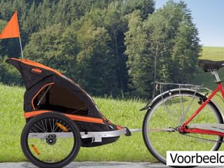 Rode fiets met zwart-oranje kinderaanhanger op gras voor bosbachtergrond. Oranje veiligheidsvlag zichtbaar op aanhanger.