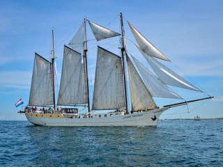 Driemast zeilschip Mare Frisium met witte zeilen op kalm blauw water onder heldere hemel, Nederlandse vlag zichtbaar.