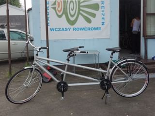 Zilveren tandemfiets met twee zitplaatsen geparkeerd voor een blauw gebouw met een fietstourenbord 'WCZASY ROWEROWE'.