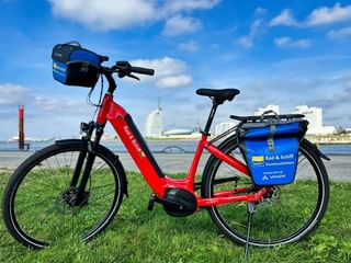 Rode SE-Tours huur e-bike met blauwe fietstassen geparkeerd op gras bij water met vuurtoren en industriële gebouwen op achtergrond.