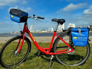 Rode SE-Tours huurfiets met blauwe Vaude tassen geparkeerd op gras bij de waterkant. Stadshorizon zichtbaar op de achtergrond.