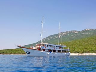Witte motoryacht MS Amore met twee masten voor anker in blauw water nabij een groen Kroatisch eiland met rotsachtige heuvels.