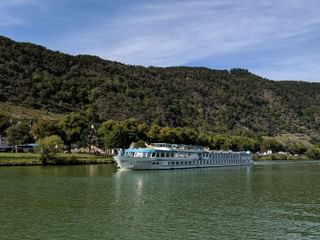 MS Switserland riviercruiseschip met witte romp en blauw bovendek varend op kalm groen water met beboste heuvels en dorp op de achtergrond.