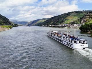 Riviercruiseschip MS Veloria varend op een brede rivier tussen groene heuvels met wijngaarden en kleine dorpen onder een gedeeltelijk bewolkte hemel.