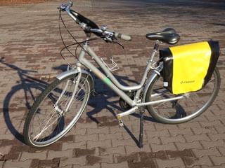 Zilveren Bird toerfiets met geel-zwarte fietstassen op achterdrager, geparkeerd op klinkers met standaard uitgeklapt.
