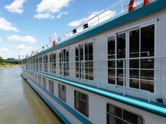 Wit MS Switzerland riviercruiseschip met blauwe rand en grote ramen, afgemeerd aan een kalme rivier onder blauwe lucht.