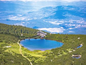 Luchtfoto van het heldere blauwe bergmeer Polezhan omringd door groene weiden en bos, met een berghut nabij en gelaagde bergen op de achtergrond.