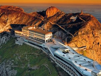 Modern bergstation gebouw op de Pilatus met dramatische rotspieken in gouden zonsondergangslicht, uitkijkend over Zwitserse valleien.