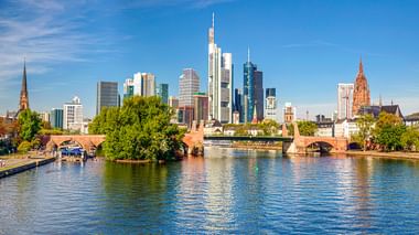 Frankfurt skyline met moderne wolkenkrabbers en historische kerktorens langs de Main. Stenen brug over het water onder blauwe lucht.