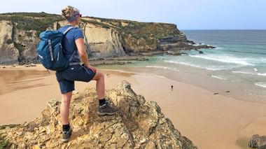 Vrouwelijke wandelaar met blauwe rugzak staat op rotsachtige uitstulping met uitzicht op zandstrand en kliffen langs de Rota Vicentina.