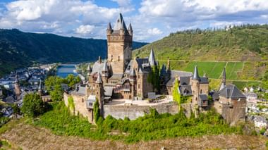 Luchtfoto van kasteel Reichsburg Cochem met torens en muren op een heuvel, omgeven door wijngaarden en de stad Cochem langs de Moezel.