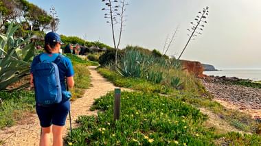 Wandelaar met blauwe rugzak op zandpad langs Portugese kust met agaveplanten en rode kliffen bij de oceaan.